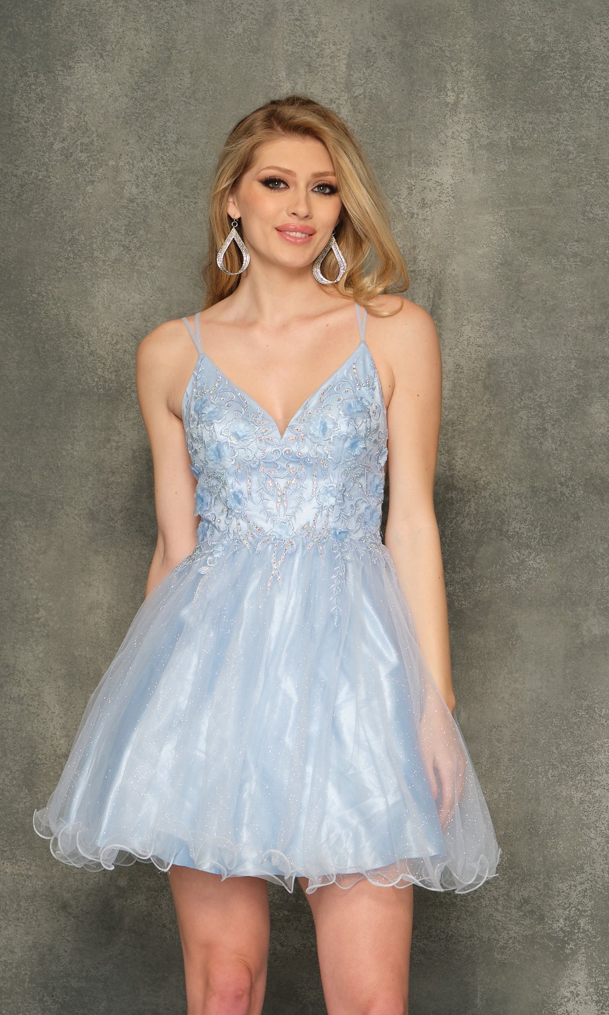 A woman wears the Sheer-Bodice Ice Blue Short Homecoming Dress 10844, a sleeveless, V-neck, fit-and-flare party dress with floral embroidery, standing against a textured gray background.
